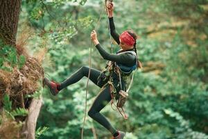 View from the side, hanging on, th,e rope. Woman is doing climbing in the forest by the use of safety equipment photo