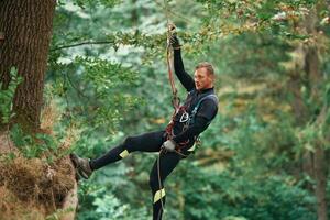 Up on the rope, hanging. Man is doing climbing in the forest by use of safety equipment photo