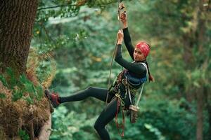 Looking to, the side, hanging on, th,e rope. Woman is doing climbing in the forest by the use of safety equipment photo