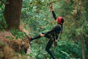View from, the side, hanging on the rope. Woman is doing climbing in the forest by the use of safety equipment photo