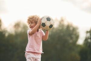 Holding,, soccer ball in hands. Cute little girl with curly hair is playing on the field at daytime photo