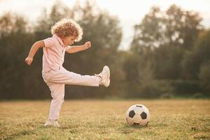 Kicks socce,r ball. Cute little girl with curly hair is playing on the field at daytime photo