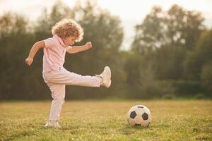 Kicks, soccer ball. Cute little girl with curly hair is playing on the field at daytime photo