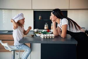 Girl is in the chief hat.. Mother with her daughter are preparing food on the kitchen photo
