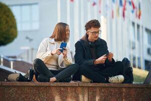 Front view. Two young students are sitting outdoors against university photo