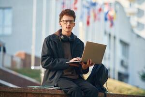 Guy is sitting and using ,laptop... Young student is outside the university, in black clothes photo