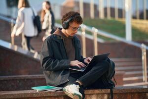 Guy is sitting and, using ,laptop. Young student is outside the university, in black clothes photo