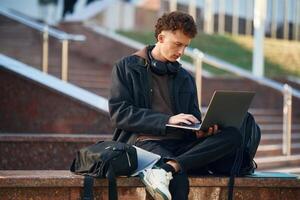 Guy, is sitting ,and using laptop. Young student is outside the university, in black clothes photo