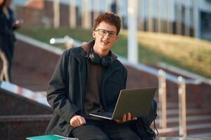 Smiling, holding laptop. Young student is outside the university, in black clothes photo