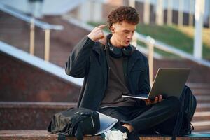 Guy, is, sitting and using laptop. Young student is outside the university, in black clothes photo