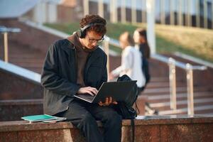Guy is, sitting and using laptop. Young student is outside the university, in black clothes photo