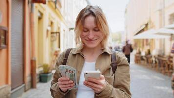 Woman counting money dollar cash using smartphone calculator app satisfied of salary on city street video