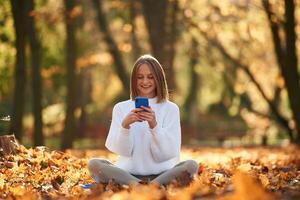 Woman, is ,sitting in the autumn park or forest with smartphone in hands photo