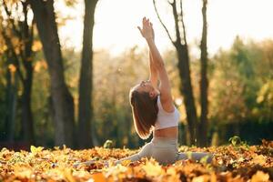Trees, at background, doing exercises. Young beautiful woman is doing yoga in the autumn park photo