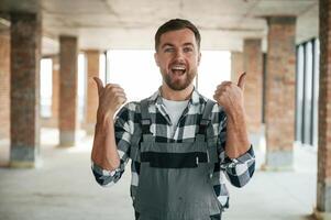With thumbs up, smiling. Construction worker in uniform in empty unfinished room photo