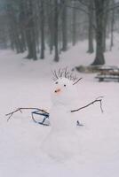 Snowman with stick hair and a carrot nose stands near a sled in a snowy meadow in the forest photo