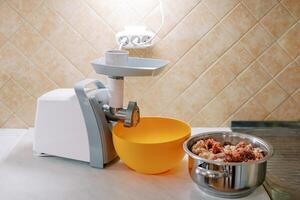 Electric meat grinder stands on a table next to a bowl and a pot of minced meat photo