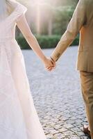 Bride and groom walk holding hands along the paving stones in the park. Back view. Cropped photo