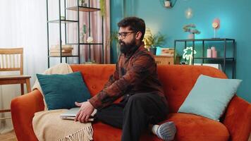 Happy young Indian man typing working on laptop browsing internet using apps sitting on home sofa video