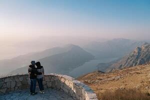Couple of travelers in jackets hug on an observation deck over the Bay of Kotor in the fog. Montenegro. Back view photo