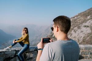 Man takes a picture on a smartphone of woman sitting on a fence in the mountains. Back view photo