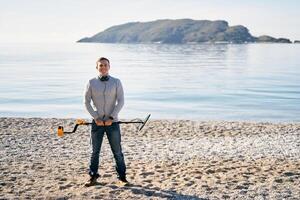 Smiling young man standing with a metal detector in his hands in front of him on the beach photo