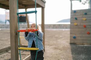Little girl stands near a rope ladder on a playground and looks up photo