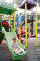 Little girl sits on a colorful slide at the playground and plays with a plush bunny photo