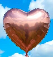 Metallic gold heart-shaped balloon floats against a bright blue sky with scattered clouds photo
