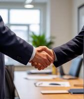 Two individuals wearing formal business suits are shaking hands across a conference table. photo
