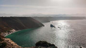 Coastal cliffs and shimmering sea under a soft hazy sky, with dramatic rock formations and distant mountains. photo