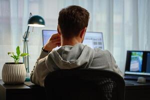 Man in gray hoodie working at computer desk with dual monitors and lamp in background. Freelancer working remotely at home office. Concept of remote work and productivity photo