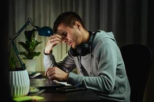 Tired man rubs eyes while working at computer. Fatigued freelancer working late at home office. Concept of stress and mental fatigue during remote work photo