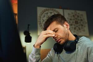 Tired man rubs eyes while working at computer. Fatigued freelancer working late at home office. Concept of mental fatigue during remote work photo