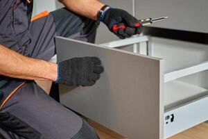 Man assembling kitchen drawer in modern cabinet using screwdriver. Handyman installing and adjusting drawer slide mechanism in contemporary kitchen. Furniture installation photo