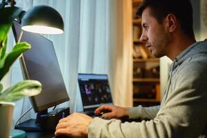 Focused man sitting by table and looking at computer monitor. Concentrated freelancer using desktop and laptop by window. Remote work from home office photo