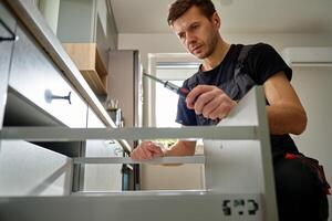 Man assembling kitchen drawer, using screwdriver. Handyman installing and adjusting drawer slide mechanism in contemporary kitchen. Furniture installation photo