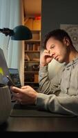 Tired freelancer procrastinating at home office, scrolling phone instead of working. Unproductive man using smartphone at home desk. Concept of procrastination and distraction video