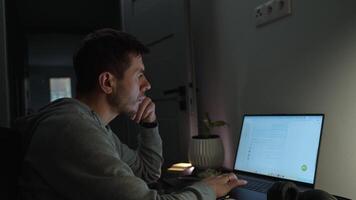 Concentrated freelancer working late at home office. Focused man in hoodie writes notes while working on laptop video