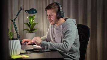 Freelancer wearing headphones working at computer in home office. Man in gray hoodie working at computer desk with dual monitors and lamp in background. Concept of remote work and productivity video