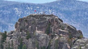 Stunning view of a rocky summit with a flag in the distance surrounded by pine trees during a clear day in the mountains video