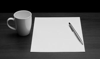 A white cup and pen on a table next to a blank sheet of paper photo