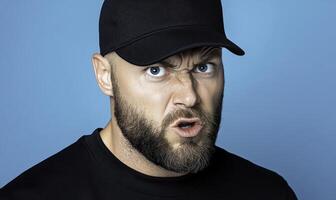 Intense Man with Beard and Hat Displaying Anger in Front of Blue Background for Diverse Emotions and Expressions in Stock Photography Needs photo