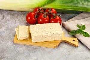 A piece of tofu on a cutting board next to a tomato and a green leaf photo