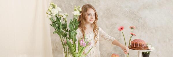 girl with long hair in a light dress is sitting at the Easter table with cakes, spring flowers and quail eggs. Happy Easter celebration. photo