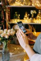 Close-up of person photographing bouquet of white tulips using smartphone in floral arrangement shop. Background features beautiful lighting and floral decor, creating warm and inviting atmosphere. photo