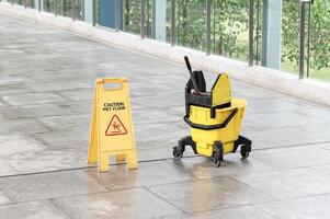 Yellow warning sign and message Caution wet floor with cleaning trolley on walkway photo