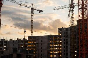 Multiple cranes work efficiently at a construction site as the sun sets, illuminating the building's framework and reflecting on workers below photo