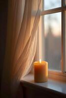 A warm, glowing candle sits on windowsill beside sheer curtains gently swaying in the soft light of dusk or dawn filtering photo