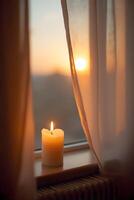 A glowing candle sits on a windowsill beside sheer curtains gently swaying in the soft light of dusk or dawn filtering photo